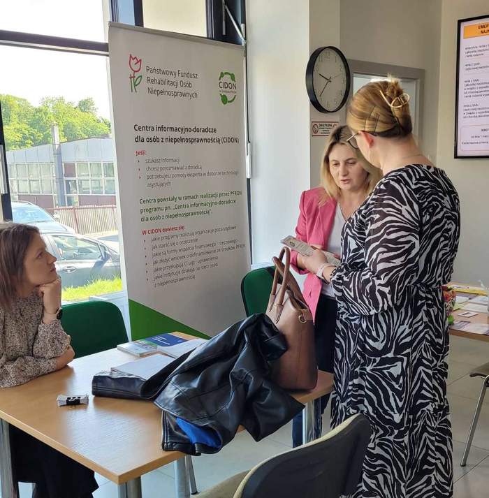 Pokaż zdjęcie: Przy stoliku siedzi jena kobieta obok niej stoją dwie kolejne i rozmawiają. W tle stoi rollup z logo CIDON, na stole leżą ulotki.