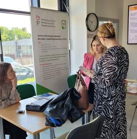 Pokaż zdjęcie: Przy stoliku siedzi jena kobieta obok niej stoją dwie kolejne i rozmawiają. W tle stoi rollup z logo CIDON, na stole leżą ulotki.