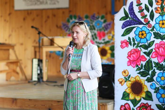 Pokaż zdjęcie: Na scenie przy mikrofonie stoi kobieta w kolorowej sukience i białym żakiecie.