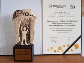 Statuetka dziękczynna dla PFRON w stylistyce logo PSONI człowiek z uniesionymi rękami na tle otwartych drzwi nad nim słońce z napisem 40 lat, pod nim napis w tym pełna nazwa koła daty 1983-2023 z prawej strony list, podziękowanie
