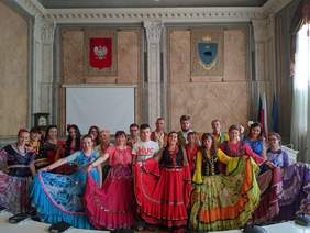 Zdjęcie w urzędzie, artyści w stojach, panie w pięknych sukniach za nimi stoją panowie