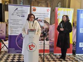 Pokaż zdjęcie: W kościele stoją dwie kobiety. Jedna z nich przemawia do mikrofonu i składa zebranym świąteczne życzenia. Panie są elegancko ubrane. 