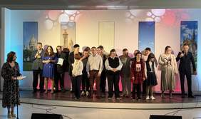 Pokaż zdjęcie: Grupa około dwudziestu osób znajduje się na dużej scenie. Z przodu stoi kobieta, która poprzez mikrofon zwraca się do zgromadzonych osób. W tle, na ścianie wyświetlane są zdjęcia prezentujące różnego rodzaju budowle z całego świata.