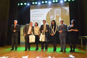 Pokaż zdjęcie: na scenie stoi w rzędzie 7 osób w tym dwaj mężczyźni trzymają w rękach nagrody w tle widać nagrodzoną pracę