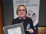 Kobieta w okularach i ciemnej marynarce trzyma oprawiony certyfikat oraz statuetkę Lodołamaczy. W tle widoczny jest napis „Zostań Lodołamaczem” oraz grafika przedstawiająca statuetkę.  