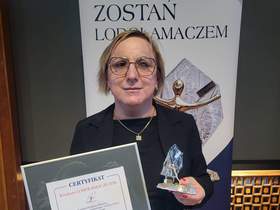 Kobieta w okularach i ciemnej marynarce trzyma oprawiony certyfikat oraz statuetkę Lodołamaczy. W tle widoczny jest napis „Zostań Lodołamaczem” oraz grafika przedstawiająca statuetkę.  