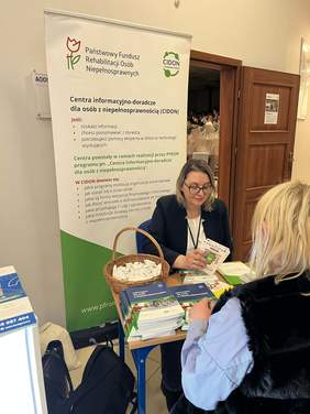 Dwie kobiety, siedzą na przeciwko siebie, rozmawiają, w tle biało-zielony baner PFRON