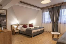 Pokaż zdjęcie: Pokój nr 11 piętro 1. Na wprost w skosie duże łóżko małżeńskie z białą pościelą nakryte szarym kocem oraz ręczniki. Po prawej stronie dwa okna z białymi firanami oraz szarymi zasłonami. Podłoga oraz wezgłowie łóżka w kolorze jasnego drewna.