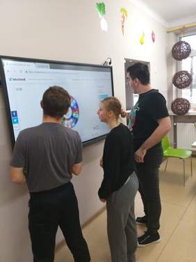 Pokaż zdjęcie: trzy osoby stoją przytelebimie i oglądają tablicę multimedialną podczas warsztatów informatycznych 