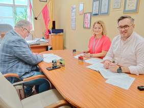 Pokaż zdjęcie: Przy podłużnym stole siedzą trzy osoby: kobieta i dwóch mężczyzn. Podpisują umowę. Na stole leżą różne dokumenty, pieczątki, telefon, długopis. 