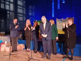 Pokaż zdjęcie: Na scenie stoją zaproszeni goście. Są to kobiety i mężczyźni. Wszyscy mają na sobie eleganckie i wytworne ubrania. 