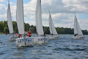 Pokaż zdjęcie: Pięć łodzi żegluje po jeziorze. Na łodziach znajdują się ludzie w pomarańczowych kapokach.