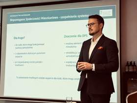 Dyrektor Oddziału Zachodniopomorskiego PFRON podczas wystąpienia na konferencji. Mężczyzna stoi przed ekranem prezentacyjnym, na którym wyświetlona jest prezentacja dotycząca wspomaganych społeczności mieszkaniowych i ich znaczenia dla domów pomocy społecznej.