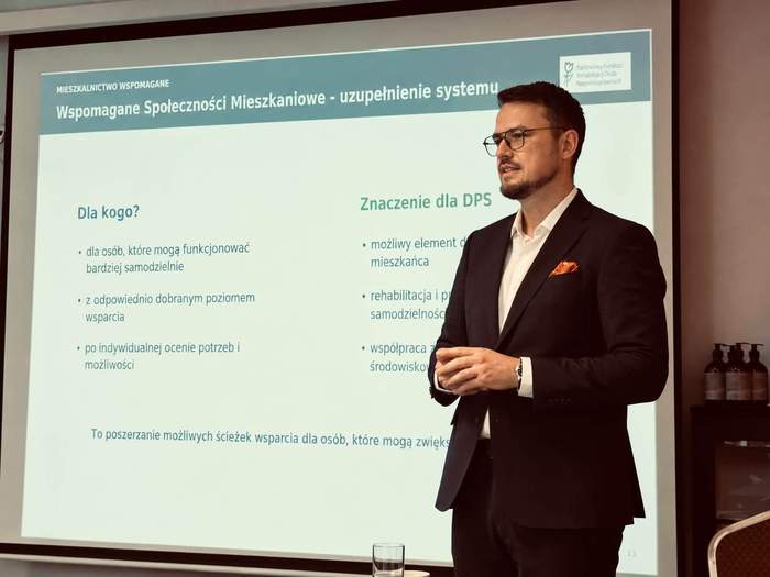 Dyrektor Oddziału Zachodniopomorskiego PFRON podczas wystąpienia na konferencji. Mężczyzna stoi przed ekranem prezentacyjnym, na którym wyświetlona jest prezentacja dotycząca wspomaganych społeczności mieszkaniowych i ich znaczenia dla domów pomocy społecznej.