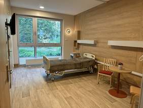Pokaż zdjęcie: Pomieszczenie z kaflami i płytami we wzorze drewna na podłodze i ścianach. Na środku łóżko rehabilitacyjne krzesło stolik. Po lewej stronie wisi telewizor naprzeciw drzwi przy łóżku okno na cała ścianę z widokiem na las.