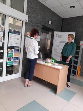 Pokaż zdjęcie: Przy biurku na którym leżą ulotki rozmawiają dwie kobiety, w tle baner z logo PFRON.