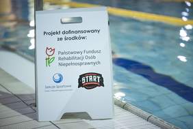 Pokaż zdjęcie: Tablica informacyjna z napisem Projekt dofinansowany ze środków: Państwowy Funduszu Rehabilitacji Osób Niepełnosprawnych oraz Sekcje Sportowe Osób z Niepełnosprawnościami oraz logo Stowarzyszenia START Białystok. W tle rozmazany obraz wody w basenie.