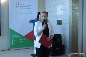 Pokaż zdjęcie: Kobieta blondynka w okularach załozonych na włosy, stoi przed gośćmi zaproszonymi na konferencję, w jednej ręce trzyma mikrofon w drugiej dokumenty
