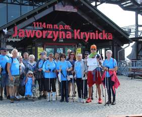Pokaż zdjęcie: Grupa osób w różnym wieku stoi przed wejściem do kolejki gondolowej na Jaworzynę Krynicką. Prawie wszystkie osoby są ubrane w błękitne koszulki i trzymają w dłoniach kijki. 