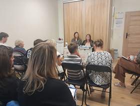 Spotkanie grupy osób w niewielkiej sali. Uczestnicy siedzą na krzesłach ustawionych w rzędach i słuchają dwóch kobiet siedzących przy stole z materiałami informacyjnymi.