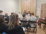 Spotkanie grupy osób w niewielkiej sali. Uczestnicy siedzą na krzesłach ustawionych w rzędach i słuchają dwóch kobiet siedzących przy stole z materiałami informacyjnymi.