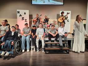 Pokaż zdjęcie: Na scenie znajdują się dzieci. Jedne siedzą, a inne stoją. Mają otwarte buzie i śpiewają piosenkę. Jedna dziewczynka trzyma w dłoniach mikrofon. 