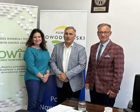 Pokaż zdjęcie: Dwaj mężczyźni i kobieta stoją na tle banerów Powiatu nowodworskiego i OWIT. Po prawej na ścianie wisi mosiężny orzeł. Przed nimi stoi biurko na którym leżą dokumenty, woda mineralna.
