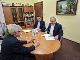 Pokaż zdjęcie: Przy owalnym stole siedzi trzech mężczyzn. Podpisują umowę. Na stole leżą różne dokumenty, pieczątki, długopisy, woda mineralna. 