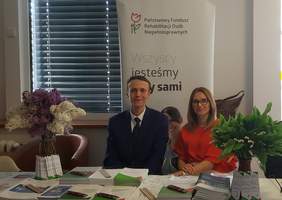Pokaż zdjęcie: Kobieta ubrana w czerwoną bluzkę i mężczyzna w granatowym garniturze siedzą na tle banera PFRON. Na stoliku stoją dwa bukiety kwiatów, tj. bzów oraz konwalii. Ponadto ułożone zostały na nim ulotki i materiały informacyjne oraz długopisy i ołówki. 