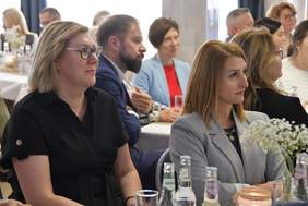 Pokaż zdjęcie: Grupa kilkunastu osób siedzi przy stolikach, kierując swój wzrok w jedną stronę. Na stolikach znajdują się bukiety białych, drobnych kwiatów, szklane butelki z wodą mineralną, jak również szklanki.