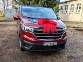 Pokaż zdjęcie: Mikrobus w kolorze bordowym z czerwoną kokardą na masce pojazdu. W tle ciąg niskich zabudowań, blaszany garaż, drzewa oraz bezchmurne niebo.