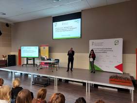 Pokaż zdjęcie: duża sala konferencyjna po środku wisi duży telebim z informacją o programie CIDON na platformie stoją dwie osoby i przemawiają do publiczności