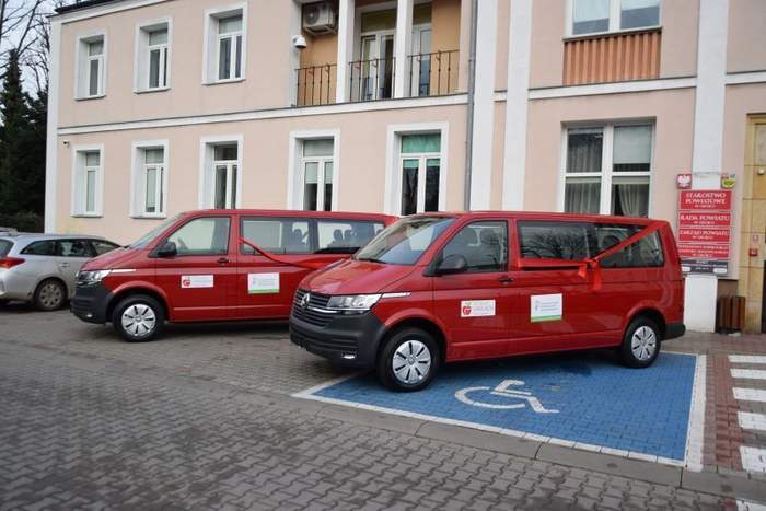 Pokaż zdjęcie: Dwa samochody (busy 9-cio miejscowe) stojące przed budynkiem w którym mieści się siedziba Starostwa Powiatowego w Grójcu. Samochody są koloru wiśniowego, na bokach naklejona m.in. naklejka iformująca o dofinansowaniu zakupu ze środków PFRON.Samochody przepasane czerwoną wstęgą. 