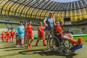 Osoby uczestniczące w turnieju piłkarskim na murawie Polsat Plus Areny Gdańsk. Na pierwszym planie osoba na wózku inwalidzkim w czerwonym stroju sportowym, obok opiekun i grupa zawodników. W tle widoczne trybuny stadionu i boisko.