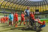 Osoby uczestniczące w turnieju piłkarskim na murawie Polsat Plus Areny Gdańsk. Na pierwszym planie osoba na wózku inwalidzkim w czerwonym stroju sportowym, obok opiekun i grupa zawodników. W tle widoczne trybuny stadionu i boisko.