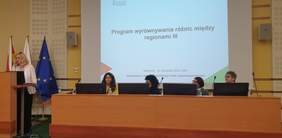 Pokaż zdjęcie: Na zdjęciu sala konferencyjna, w której odbywa się prezentacja na temat „Program wyrównywania różnic między regionami III”. Na pierwszym planie stoi kobieta w jasnej marynarce, przemawiająca przy mównicy z mikrofonem i laptopem. Za nią widać flagi Polski, Unii Europejskiej oraz inny sztandar. Obok mównicy znajduje się długi stół, przy którym siedzą cztery kobiety, każda przy własnym monitorze i mikrofonie. W tle wyświetlona jest prezentacja z datą Białystok, 15 listopada 2024 roku oraz nazwą organizatora: Państwowy Fundusz Rehabilitacji Osób Niepełnosprawnych.