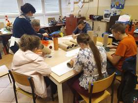 Pokaż zdjęcie: Przy ławie szkolnej siedzi pięcioro uczniów i robią prace plastyczne, wspomaga ich nauczycielka, stojąca z boku