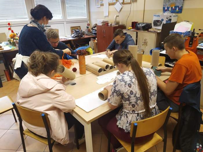 Pokaż zdjęcie: Przy ławie szkolnej siedzi pięcioro uczniów i robią prace plastyczne, wspomaga ich nauczycielka, stojąca z boku