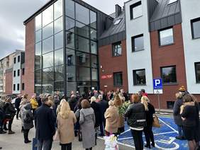 Pokaż zdjęcie: Nowy budynek z elewacją pół na pół cegła i biała ściana na zmianę. Po środku monumentalna szklana klatka schodowa z windą w środku. Przed nim wielu ludzi z kwiatami ubranych na galowo w płaszczach.