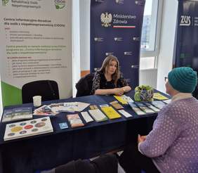 Pokaż zdjęcie: Przy stoliku na którym leżą ulotki i foldery siedzą dwie Panie. Jedna z pań udziela informacji drugiej. W tle rollup z logo Funduszu.