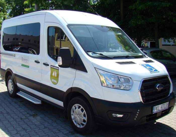 Pokaż zdjęcie: Biały bus z logo PFRON i Gminy Kościelec oraz niebieską naklejką informującą, że pojazd przystosowany jest do przewozu osób niepełnosprawnych, w tym poruszających się na wózku inwalidzkim. W tle widoczny jest fragment budynku, samochód osobowy, jak również alejka drzew liściastych.