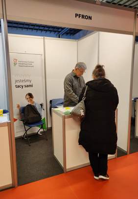 Pokaż zdjęcie: Stoisko Państwowego Funduszu Rehabilitacji Osób Niepełnosprawnych na Targach Pracy w Białymstoku. Przedstawiciel PFRON, starszy mężczyzna w okularach i szarej koszuli, rozmawia z kobietą ubraną w czarną kurtkę. Na stoliku znajdują się ulotki oraz materiały informacyjne. W tle widoczny jest baner z logo PFRON i hasłem „Wszyscy jesteśmy tacy sami”.