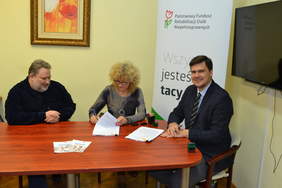 Pokaż zdjęcie: Trzy osoby siedzą przy kwadratowym stole. Mężczyzna z lewej patrzy w stronę kobiety podpisującej kolejne strony umowy. Mężczyzna z prawej uśmiecha się do obiektywu.