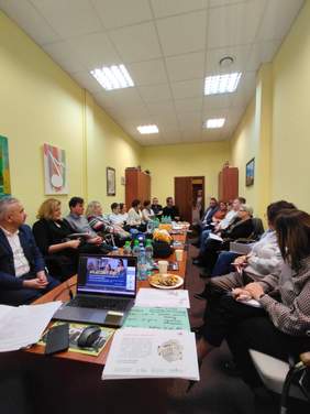 Pokaż zdjęcie: W sali konferencyjnej przy podłużnym stole siedzi kilkanaście osób. Rozmawiają ze sobą. Na stole leży komputer z wyślietloną prezentacją, notesy, skoroszyty, napoje i ciastka. 