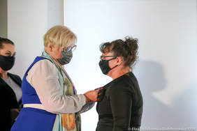 Pokaż zdjęcie: Kobieta w granatowej sukience i z kolorowym szalem przypina odznaczenie do marynarki kobiety w czarnej maseczce i garsonce.