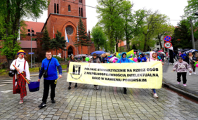Uczestnicy przemarszu idący brukowaną ulicą przed dużą, ceglaną katedrą w Kamieniu Pomorskim. Kilka osób niesie duży żółty transparent z czarnym napisem: „POLSKIE STOWARZYSZENIE NA RZECZ OSÓB Z NIEPEŁNOSPRAWNOŚCIĄ INTELEKTUALNĄ KOŁO W KAMIENIU POMORSKIM”. Po lewej stronie idzie klaun w charakterystycznym stroju, a za transparentem widać tłum ludzi z kolorowymi parasolami i balonami.