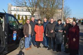 Pokaż zdjęcie: Na zdjęciu grupa osób stojąca przy czarnym busie na tle budynków i drzew. Po lewej stronie widoczna część pojazdu z białym napisem „GMINA” i niebiesko-białym herbem. W środku osiem osób ustawionych w rzędzie: kilku mężczyzn i kilka kobiet w zimowych. Zdjęcie wykonane podczas uroczystości przekazania busa do przewozu osób z niepełnosprawnościami.