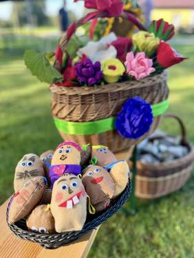 Pokaż zdjęcie: Maskotki w kształcie ziemniaka z oczami, zębami i dużym uśmiechem, w tle koszt z kwiatami. 