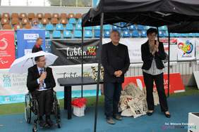 Pokaż zdjęcie: Na stadionie, pod namiotem, stoja trzy osoby, mężczyzna w garniturze na wózku, przemawia do mikrofonu, dalej mężczyzna w garniturze oraz kobieta w spodniach i marynarce.