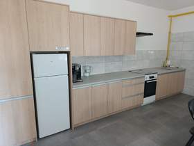 Wyremontowana kuchnia w jasnym drewnie z długim, prostym blatem. Po lewej stronie widać wysoką zabudowę i białą lodówkę z dwoma drzwiami (zamrażalnik u góry, chłodziarka na dole). Nad blatem są szafki wiszące w tym samym jasnym drewnie. Między blatem a szafkami są jasnoszary płytki. Na blacie, obok lodówki stoi czarny ekspres do kawy. Dalej w prawo jest płyta grzewcza i piekarnik (front czarny z białą ramką). Skrajnie po prawej jest zlew z kranem. Przy prawej ścianie widać żółtą rurę biegnącą pionowo i poziomo.